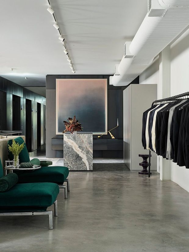 A sophisticated open-plan retail interior combining a clothing display area with a lounge-like seating zone. Dark built-in cabinetry and wall panels contrast with pale walls, polished concrete flooring, exposed white ductwork, track lighting, and a dramatic stone-clad central plinth. Deep green upholstered seating, monochromatic clothing racks, and a large abstract artwork create a refined, gallery-like atmosphere.