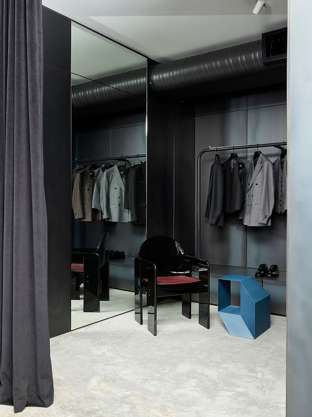 A dark, industrial-style walk-in wardrobe or dressing room with black built-in cabinetry and open clothing rails, mirrored wall panels, exposed cylindrical ductwork, and a polished concrete floor. A black armchair with a red seat cushion and a sculptural blue side table add bold colour accents. A heavy charcoal curtain frames the room.