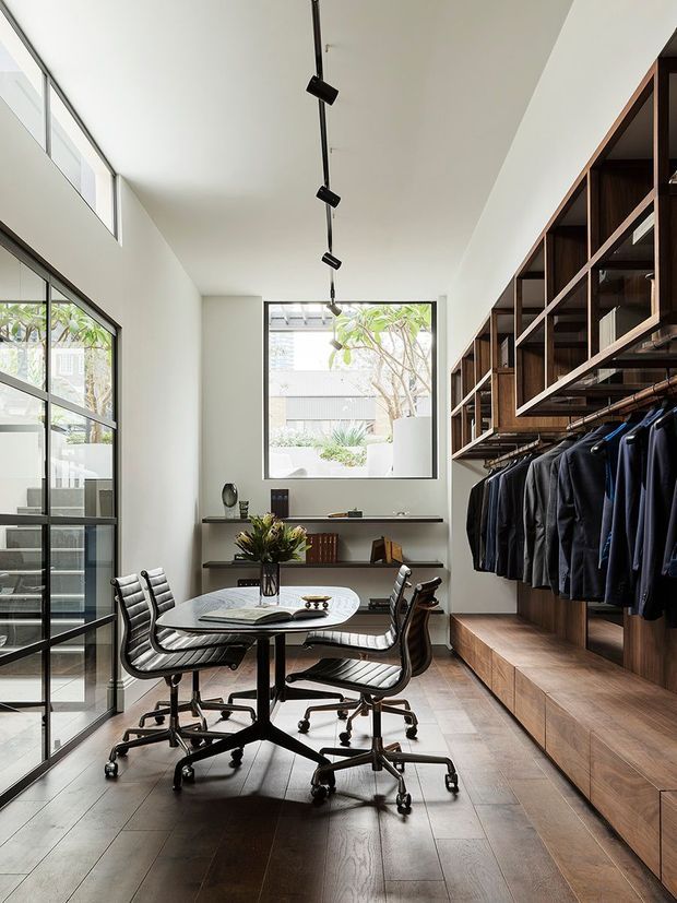 A bright, narrow contemporary dressing room or walk-in wardrobe with a built-in dark timber display and hanging system along the right wall. Multiple dark garments hang below open shelving and cabinetry, while a round meeting table with four black leather office chairs occupies the centre. Floor-to-ceiling glazed doors and windows bring in abundant daylight, and a high white ceiling with a linear track light emphasizes the room's height. The floor is finished in dark timber, with a minimalist shelving unit and decorative arrangement at the far end.