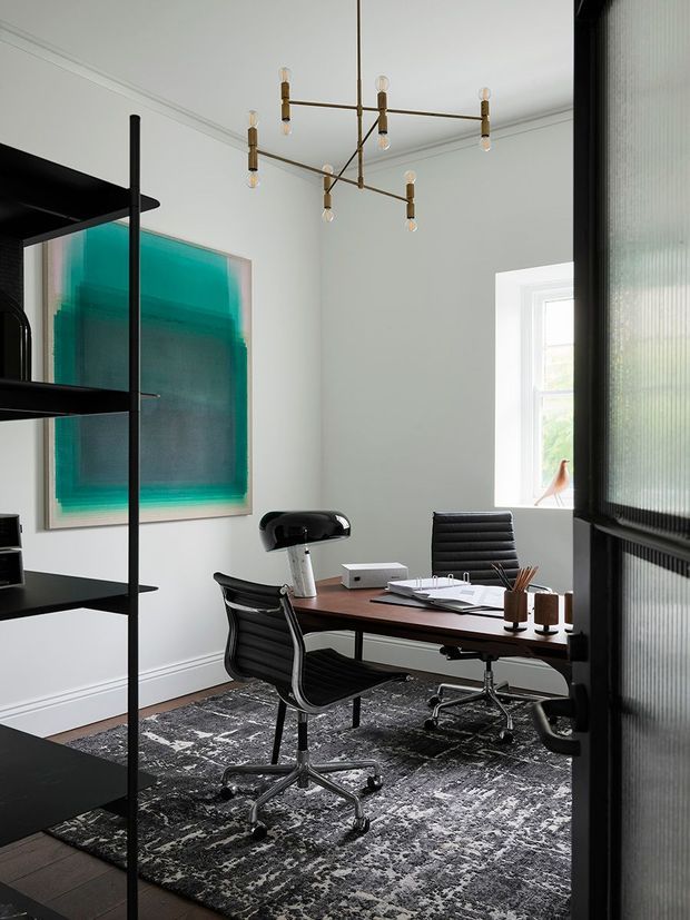 A refined home office with a dark wood desk, two black leather office chairs, open black shelving, a large abstract artwork, and a sculptural brass multi-arm pendant light. White walls with decorative cornice moulding and a patterned dark rug create a sophisticated, high-contrast workspace. A fluted translucent glass door or partition is visible on the right.