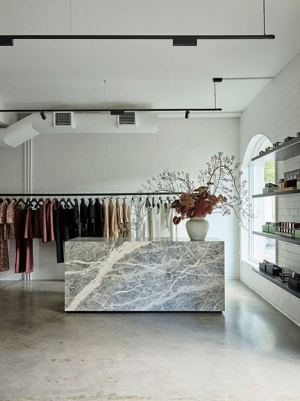 A bright, minimalist fashion retail boutique with white painted brick walls, polished concrete flooring, exposed HVAC ductwork, and black linear track lighting. A large veined stone retail counter occupies the centre, with clothing racks behind it and dark built-in display shelves along the right wall. An arched window adds natural light, while a vase of dried branches sits on the counter.