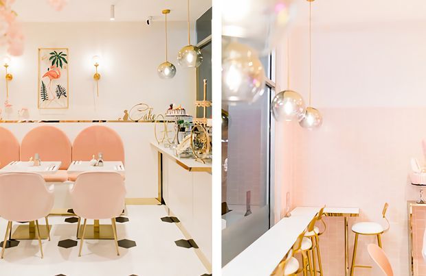 Bright pastel-toned dining and kitchen interior with blush pink upholstered dining chairs, a white dining table, pale cabinetry, a white floor with scattered black geometric inlays, and a compact breakfast counter with gold-toned stools. Multiple spherical pendant lights hang from the ceiling, while tropical and flamingo wall artwork adds playful decorative detail. A reflective glazed or mirrored wall enlarges the view of the room.