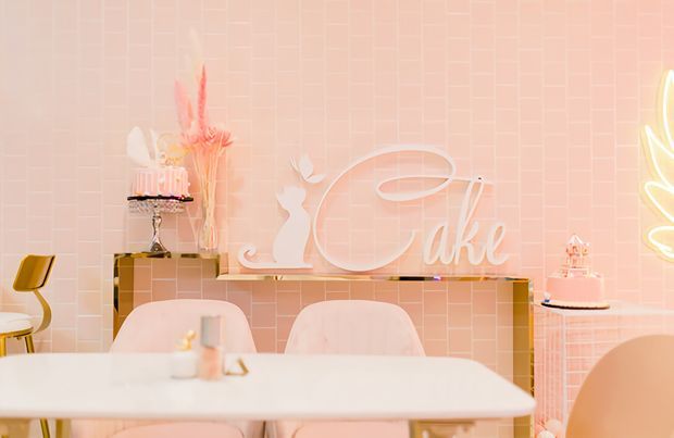 A softly lit pastel pink dining or celebration space with a white table and upholstered chairs in the foreground. The room features a pink geometric tiled wall, a slim floating console, decorative cake displays, sculptural lettering, and a bright illuminated decorative element at the right edge. The overall styling is playful, monochromatic, and refined with strong blush-pink and white tones.
