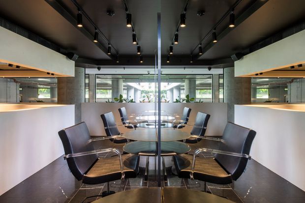 A symmetrical contemporary dining or meeting area with round tables and black leather-and-chrome chairs, viewed through a central glass partition or mirror. White curved built-in joinery or counters line both sides, with warm timber shelf accents, a dark ceiling, and exposed track lighting. Large glazed openings provide views into a bright courtyard or adjoining space.