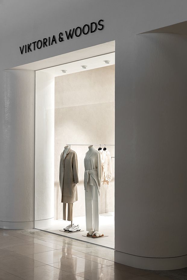 Minimalist retail clothing storefront with a recessed, brightly illuminated display alcove. Neutral white and pale beige finishes, mannequins in understated garments, integrated ceiling spotlights, curved plastered wall forms, and a highly reflective light tile floor create a refined boutique setting. The storefront signage reads “VIKTORIA & WOODS.”