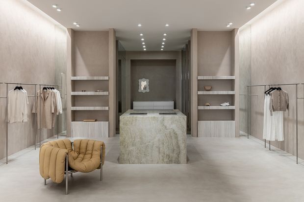 A refined, symmetrical high-end clothing boutique with pale stone-look flooring, textured neutral walls, integrated shelving, open clothing racks, and a central stone-clad display island. Soft recessed lighting, muted furnishings, and a sculptural tan lounge chair create a minimalist luxury retail interior.