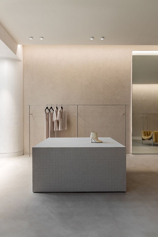 A minimalist fashion retail interior with a warm, monochromatic palette. The space features a large tiled rectangular display plinth, pale plaster walls, a smooth concrete-like floor, recessed ceiling spotlights, concealed lighting, hanging garments, and a full-height mirror reflecting seating and the display area.