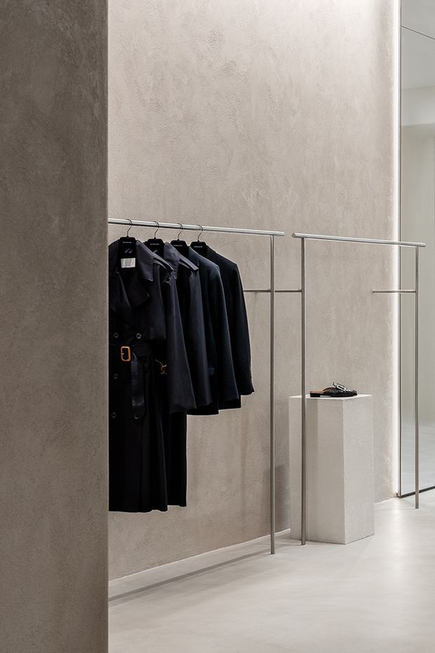 Minimalist retail clothing display with dark garments hanging from slim metal rails against textured light-grey plaster walls. A simple pale pedestal displays accessories, and a glass doorway or partition opens to a bright adjoining space. The restrained palette and clean lines create a refined, gallery-like atmosphere.