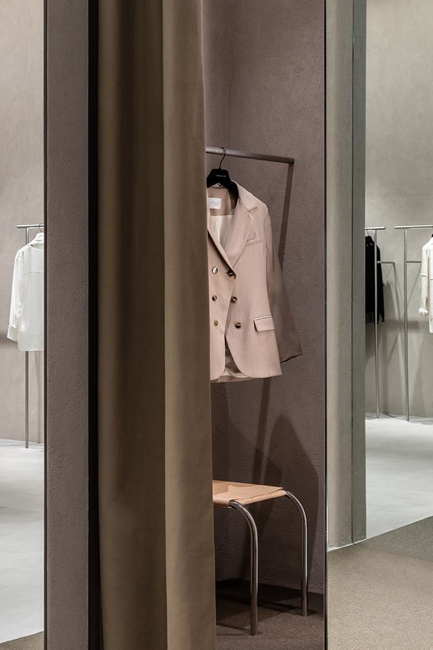 A refined clothing retail fitting area with a taupe textured partition, a full-height mirror, minimalist metal garment rails, a suspended blush-beige blazer, and a small upholstered stool. The space uses muted neutral tones, smooth concrete-like flooring, and soft contemporary lighting.