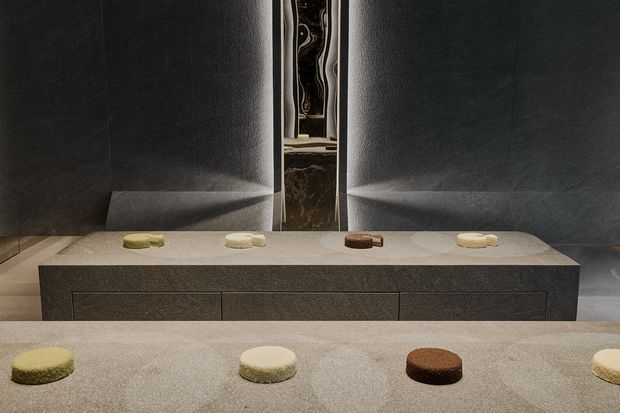 A dark, refined bathroom vanity viewed straight on, featuring a long pale stone benchtop with multiple sculptural vessel basins, dark textured wall panels, and a broad dark floating or integrated vanity with drawers. A narrow central mirror or reflective opening creates a symmetrical composition, with subtle warm neutral basin accents.