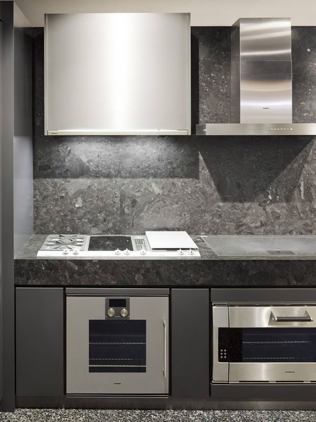 A compact contemporary kitchen with dark grey stone-look wall and benchtop surfaces, matte dark cabinetry, and integrated stainless-steel cooking appliances. The composition includes a cooktop, oven, microwave, rangehood, and sink, with a restrained monochromatic palette and metallic highlights.