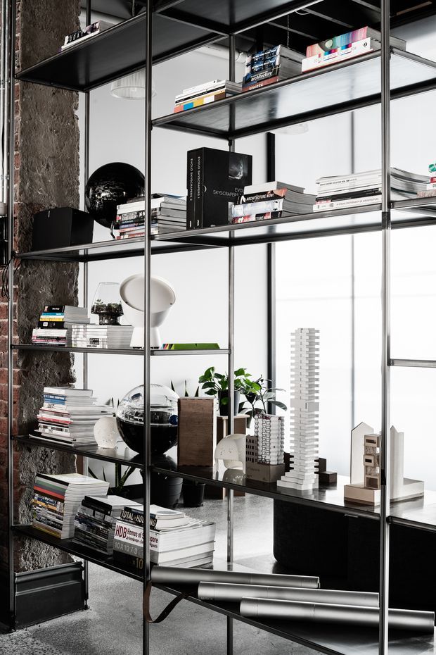 A contemporary office or studio interior featuring a tall open black metal shelving system filled with architecture and design books, objects, models, plants, and lighting. The shelving stands in front of large bright windows and beside a rough exposed concrete or masonry column. A black desk or cabinet is visible behind the shelving, with rolled drawings in the foreground. The space has an industrial, gallery-like atmosphere with strong black-and-white contrast.