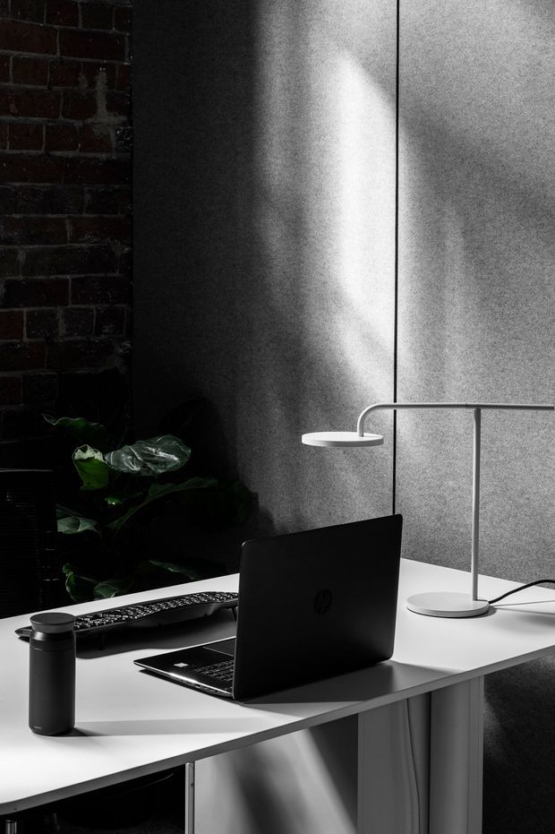 A moody, high-contrast home workspace featuring a black laptop and keyboard on a white built-in desk, with a slim curved desk lamp and a dark water bottle. The space has a dark exposed brick wall, grey textured wall panels, strong directional sunlight, and a leafy indoor plant in the corner. The monochrome palette and dramatic shadows create a refined, focused atmosphere.