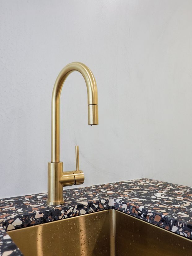Close-up product-style image of a brushed brass/gold gooseneck kitchen mixer tap mounted beside a deep undermount sink. The sink and tap are set into a dark terrazzo benchtop with cream, grey, brown and blue stone chips, against a plain light plaster wall.