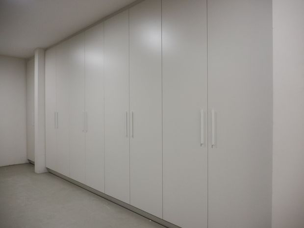 A minimalist, mostly empty room dominated by a long wall of floor-to-ceiling white built-in wardrobes with slim vertical handles. The space has plain white walls and ceiling, a pale smooth floor, and subdued lighting.