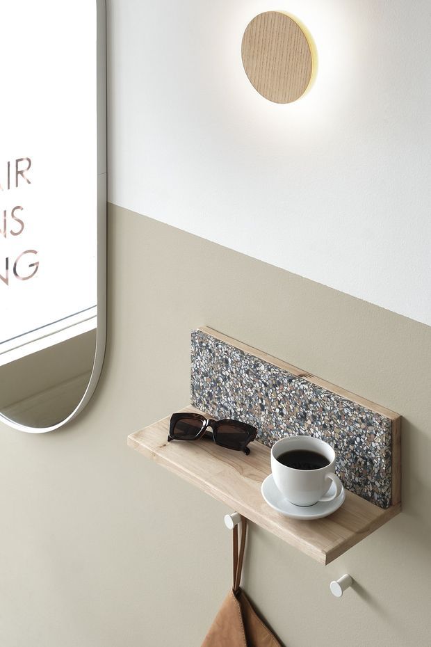 A close-up interior vignette showing a compact wall-mounted timber shelf with a terrazzo-like raised back panel, a coffee cup, sunglasses, and hooks below. The wall is painted in a soft two-tone beige and off-white diagonal composition, with a rounded-edge mirror visible at left and a circular timber wall feature above.
