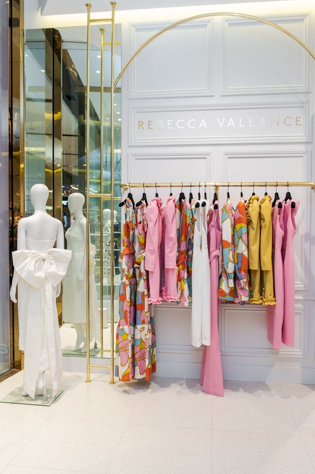 A brightly lit luxury fashion boutique interior featuring a white decorative wall with raised rectangular panelling and the “Rebecca Vallance” name, a brass/gold clothing rail displaying colourful garments, white mannequins, and mirrored glass display cabinetry. The space has a polished, refined retail fit-out with pale flooring and strong vertical reflections.