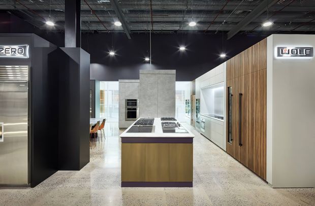 A contemporary commercial kitchen showroom with a central island, integrated cooking appliances, tall display cabinetry, timber and white wall units, polished concrete-style flooring, and an exposed dark industrial ceiling. Wolf and Sub-Zero appliance branding is visible.