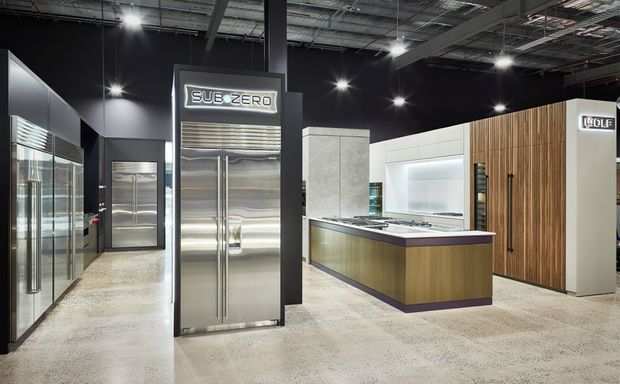 A high-end appliance showroom displaying large stainless-steel refrigeration units, including prominent Sub-Zero and Wolf appliances. The commercial interior has dark walls and exposed industrial services above, bright track and spot lighting, polished concrete flooring, and a central kitchen display with wood cabinetry, a dark benchtop, and white wall cabinetry.