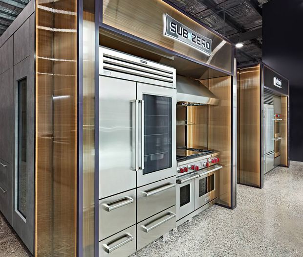 A commercial showroom display featuring high-end stainless-steel kitchen appliances, including a large refrigerator, range, and rangehood, integrated into a dark, metallic and timber-toned retail exhibition wall. The display is brightly lit with illuminated signage and set within an industrial-style showroom with an exposed ceiling.