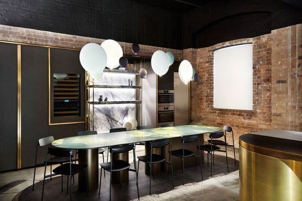 A dramatic, moody open-plan dining and entertaining space with an elongated stone dining table surrounded by black chairs. The room combines exposed brick walls, dark painted brickwork, black cabinetry with brass detailing, integrated shelving, a wine storage display, sculptural pendant lighting, and a curved brass-fronted bar or counter. A large blank projection screen or illuminated panel is set into the exposed brick wall.