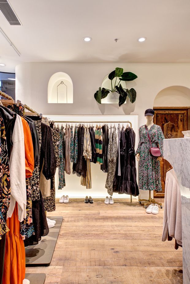 A boutique clothing retail interior with warm light timber flooring, white plaster walls, arched niches and doorways, illuminated clothing rails, mannequins, and a marble-look checkout or display counter. Decorative hanging greenery and timber architectural details add warmth to the refined shop fit-out.