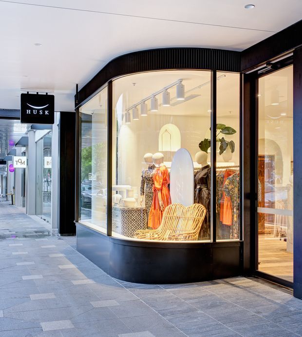 Street-level view of a contemporary fashion boutique storefront in a covered shopping arcade. The shop has a curved, floor-to-ceiling glazed display window, black framing and fascia, warm interior lighting, mannequins in colourful garments, and a woven rattan chair visible in the window display. Grey tiled paving and neighbouring retail signage surround the entrance.