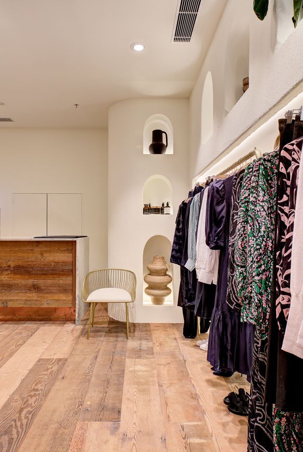 A warm, softly lit clothing boutique or walk-in wardrobe with rows of hanging garments along the right wall, a rustic timber checkout or storage counter on the left, pale timber flooring, and sculptural arched wall niches displaying ceramics and accessories. The space has curved plaster detailing, recessed lighting, and a refined earthy palette.