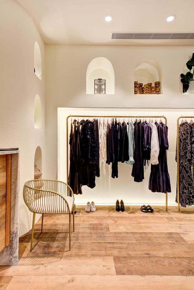 Warm, softly lit retail clothing boutique with an arched, textured plaster interior. Clothing is displayed on brass-toned garment rails within a recessed illuminated alcove, with shoes arranged below. Built-in arched niches display accessories, while a sculptural wire chair and timber flooring add to the refined, boutique atmosphere.