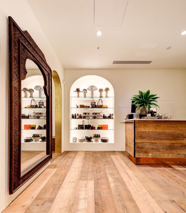 A refined retail showroom or boutique interior with warm timber flooring, cream plaster walls, arched recessed display niches, and illuminated glass shelving presenting small merchandise. A large ornate dark carved timber mirror occupies the left wall, while a substantial rustic timber reception or display counter with a stone-like top anchors the right side. Recessed ceiling lighting and a large potted plant add to the polished, welcoming fit-out.