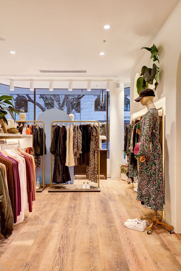 Bright contemporary clothing boutique interior with warm timber plank flooring, white walls and ceiling, recessed and track lighting, full-height glazing, and freestanding gold-toned garment racks. Clothing displays and a mannequin in a patterned dress fill the retail space.