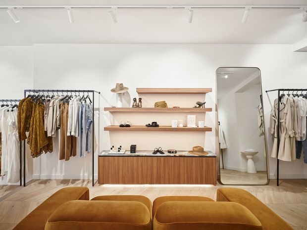 Bright contemporary clothing boutique with white walls, light timber flooring, freestanding black metal garment racks, and a central walnut-toned retail display cabinet with open timber shelves. A large rounded mirror reflects a small restroom area, while plush mustard-brown seating occupies the foreground. Track lighting provides even illumination.