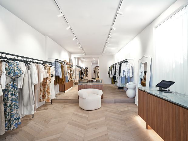 Bright contemporary clothing boutique with clothing racks arranged along both sides of a long, narrow retail space. The interior features white walls and ceiling, track lighting, pale herringbone timber flooring, warm wood retail cabinetry, curved cream seating, sculptural white stools, a full-height mirror, and a glass-topped checkout counter.