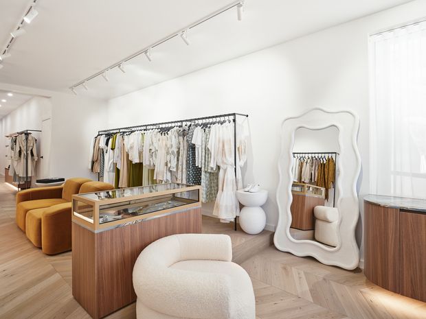 Bright, refined clothing boutique with white walls, track lighting, warm herringbone timber flooring, freestanding clothing racks, timber-and-glass display counters, sculptural upholstered seating, and a large organically shaped decorative mirror. The space has a minimalist, softly luxurious retail fit-out with warm neutral tones.