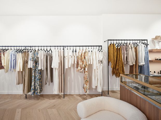 Bright contemporary clothing boutique interior with extensive garments displayed on black metal racks, a glass-topped timber display counter, shelving, and a sculptural cream boucle lounge chair. The space has white walls, light timber herringbone flooring, and a clean, refined retail fit-out.