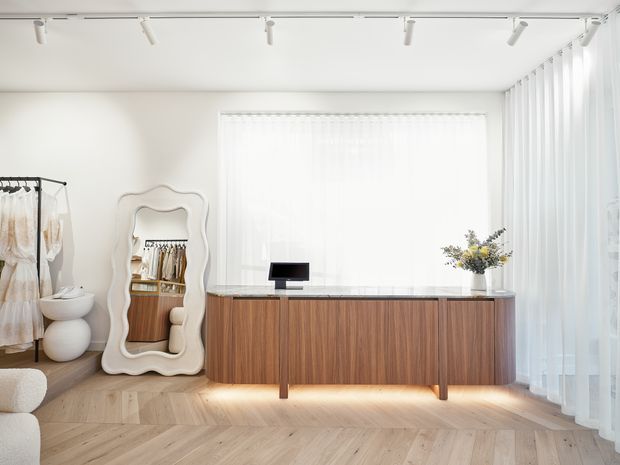 Bright, minimalist dressing and storage area with a warm timber sideboard or built-in cabinet, pale herringbone timber flooring, full-height sheer curtains, track lighting, a freestanding wavy-edged mirror, and a clothing rack visible at left. The space has a clean, refined contemporary appearance with abundant diffuse daylight.