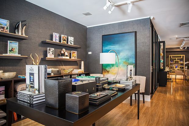 A refined, dark-toned interior featuring a styled display and work table in the foreground, wall-mounted shelves filled with books and objects, textured charcoal walls, timber flooring, track lighting, and a framed artwork. A corridor or adjoining open area with additional shelving and furnishings is visible in the background.