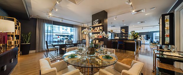 A sophisticated open-plan apartment interior combining a dining area, living space, and kitchen or bar zone. The foreground features a round glass dining table with upholstered cream chairs, while dark timber cabinetry, display shelving, pendant lighting, and a central kitchen island define the room. Floor-to-ceiling glazing and curtains bring in urban daylight, with timber flooring and a neutral palette accented by black and dark wood finishes.