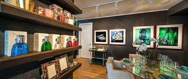 A refined dining and display area with dark textured walls, warm timber flooring, extensive floating display shelving, framed artwork, and a glass dining table set with green glassware. Decorative bottles and objects are arranged on the shelving, while an orchid and sculptural centerpiece add visual interest. Track lighting and recessed ceiling lights illuminate the space.