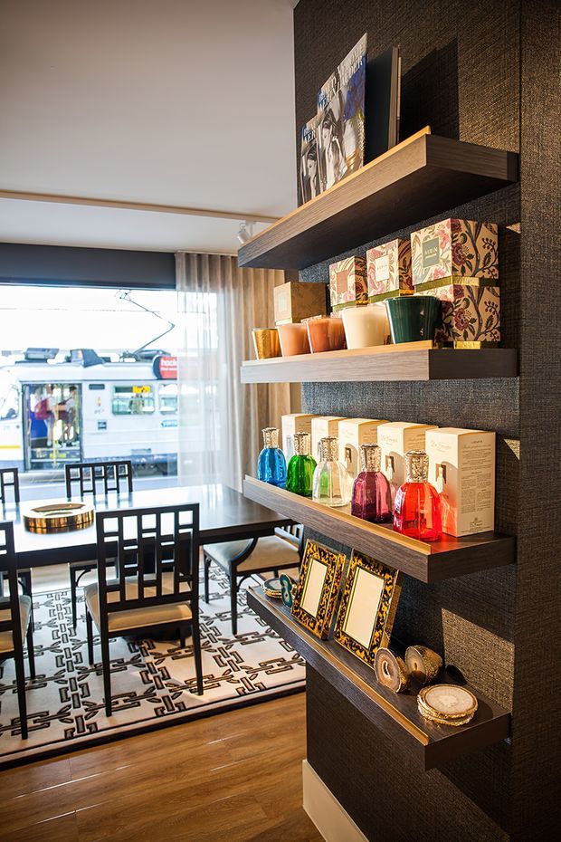 A refined dining area viewed beside a dark textured feature wall with built-in floating timber display shelves. The shelves hold colourful glass vessels, decorative boxes, framed artwork and ornaments. A black dining table with geometric-back chairs sits on a patterned monochrome rug, with timber flooring and large glazed windows opening the room to a bright urban street view. The scheme combines dark textured surfaces, warm wood and vivid decorative accents.
