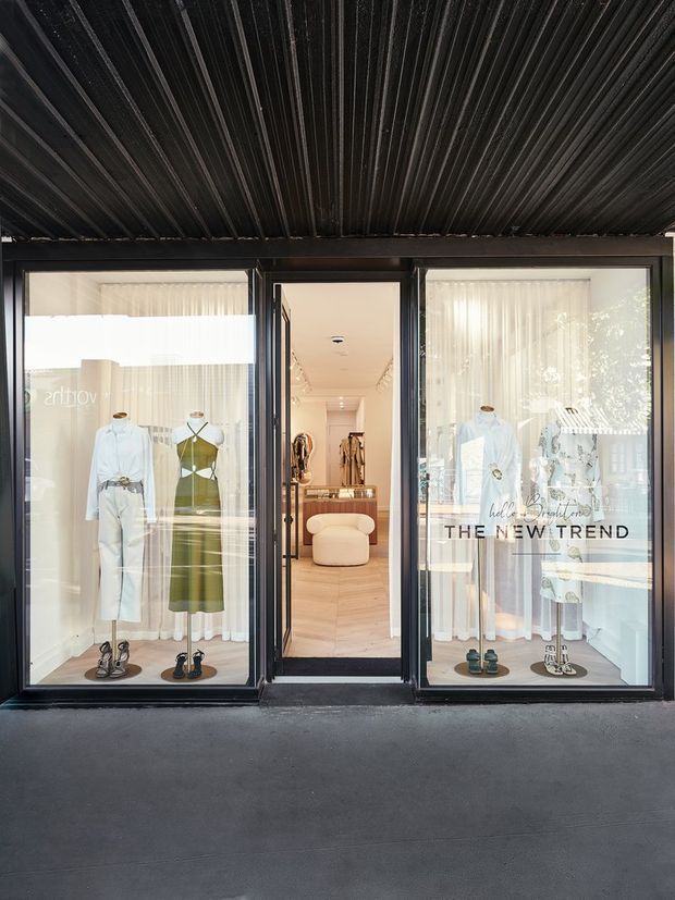 Street-facing entrance to a contemporary fashion boutique, with large black-framed glazed display windows and a central open doorway. Mannequins and clothing are visible in the window displays, while the bright minimalist retail interior beyond features pale timber flooring, clothing racks, and a rounded upholstered seating piece. A dark ribbed metal canopy spans the storefront.