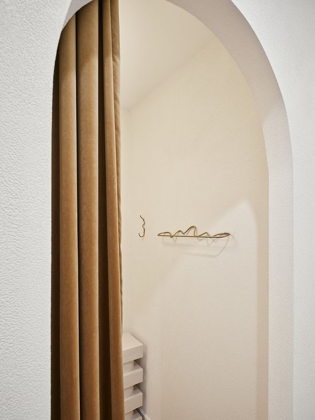 Close-up view of a softly curved, arched alcove or passage with textured off-white plaster walls. A full-height brown curtain frames the left side, while minimal wall-mounted brass or bronze hooks and a small stack of white built-in drawers are visible within the recess. The image has a warm, minimalist interior character.