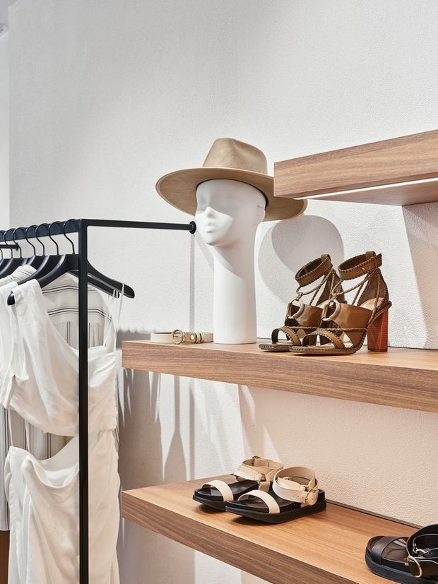 A bright, minimalist retail clothing display with black metal garment rails, white apparel, and floating warm wood shelves showcasing leather sandals, a mannequin head wearing a tan hat, and small accessories. The space has white textured walls and strong natural light creating crisp shadows.