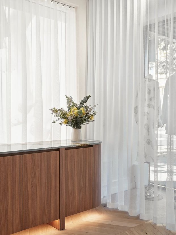 Bright, refined interior corner featuring a low dark timber credenza with a slim dark stone top and integrated underlighting. Sheer white floor-to-ceiling curtains filter abundant daylight, with a floral arrangement displayed on the cabinet and a glimpse of a bright adjacent space beyond.