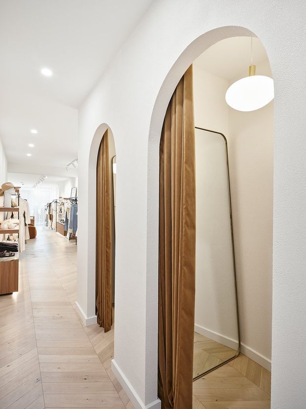 Bright, narrow retail clothing boutique interior with white textured walls, pale timber flooring, rounded arched fitting-room openings, warm wood curtains or trim, recessed ceiling lights, and a large oval pendant light. Clothing racks and shelving are visible deeper in the shop, while the foreground features a full-height mirror within an arched alcove.