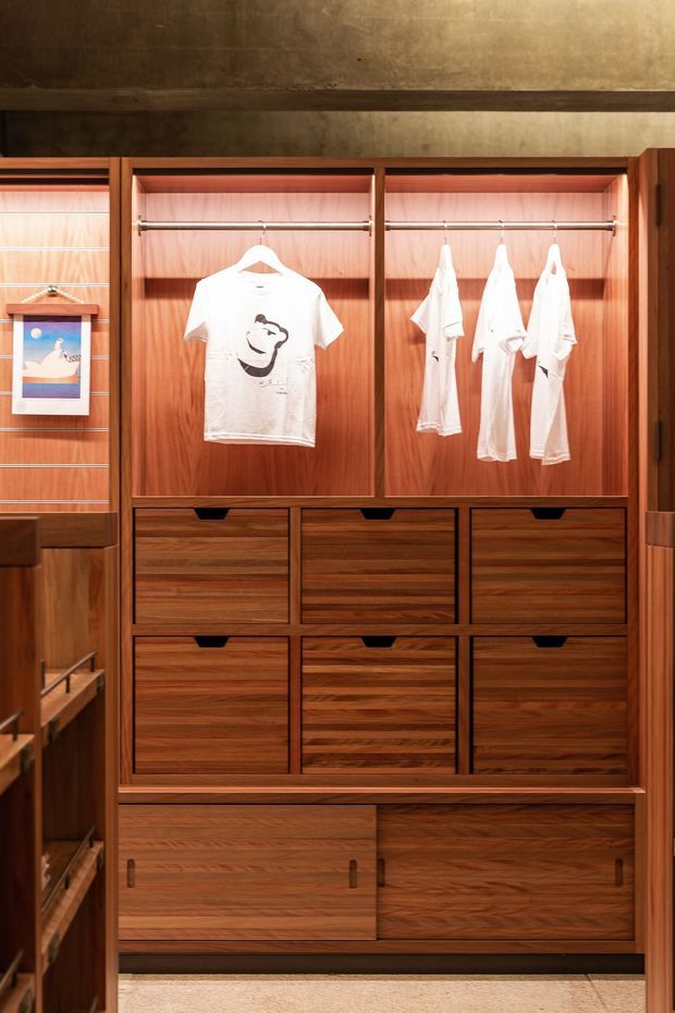 A close-up view of a custom built-in walk-in wardrobe with warm wood-grain cabinetry. The upper section has open hanging rails with white clothing, while the lower section contains multiple drawers and cupboards with recessed pulls. Integrated shelving is visible at the side, and a dark textured wall panel forms the backdrop. Warm concealed lighting highlights the timber interior.