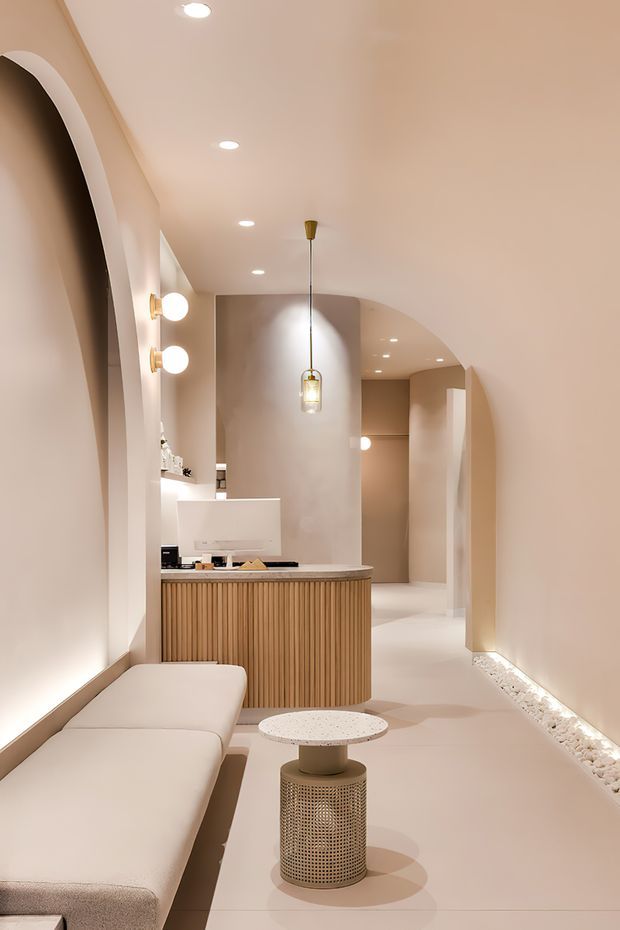 A warm, softly lit contemporary interior featuring a curved reception or concierge desk with vertical timber slat joinery, a built-in upholstered bench, a small round side table, sculptural pendant lighting, wall sconces, and a series of arched openings leading into a corridor or adjoining spaces. The palette is monochromatic cream, beige, and light timber with concealed floor-level lighting and decorative white stone detailing along the wall base.
