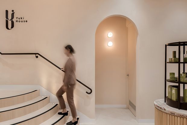 A softly lit contemporary interior stairway with curved, pale timber-clad steps, a slim dark handrail, and a blurred person ascending. An arched doorway opens to a small illuminated hall, while dark open shelving and a rounded stone-topped display counter occupy the right side. The palette is warm neutral with cream walls and light wood detailing.