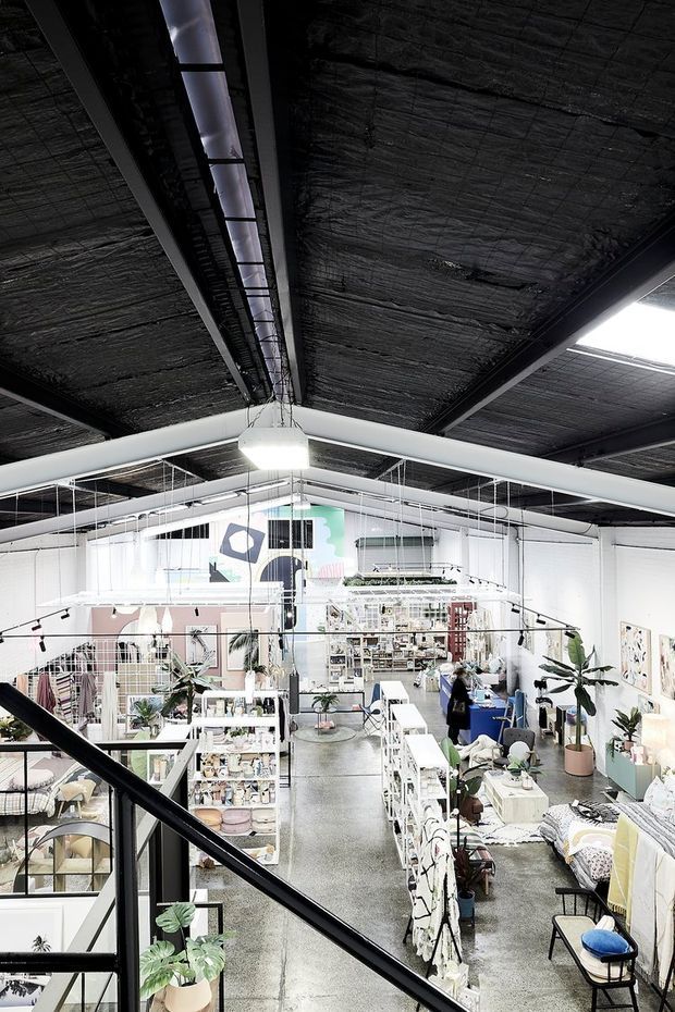 A bright, eclectic multi-level commercial studio or retail showroom viewed from an upper mezzanine. The space has exposed dark ceiling insulation and structural beams, suspended linear lighting, white walls, polished concrete flooring, extensive open shelving and display racks, plants, artwork, and a visible workspace. Black metal stair and mezzanine railings frame the foreground.
