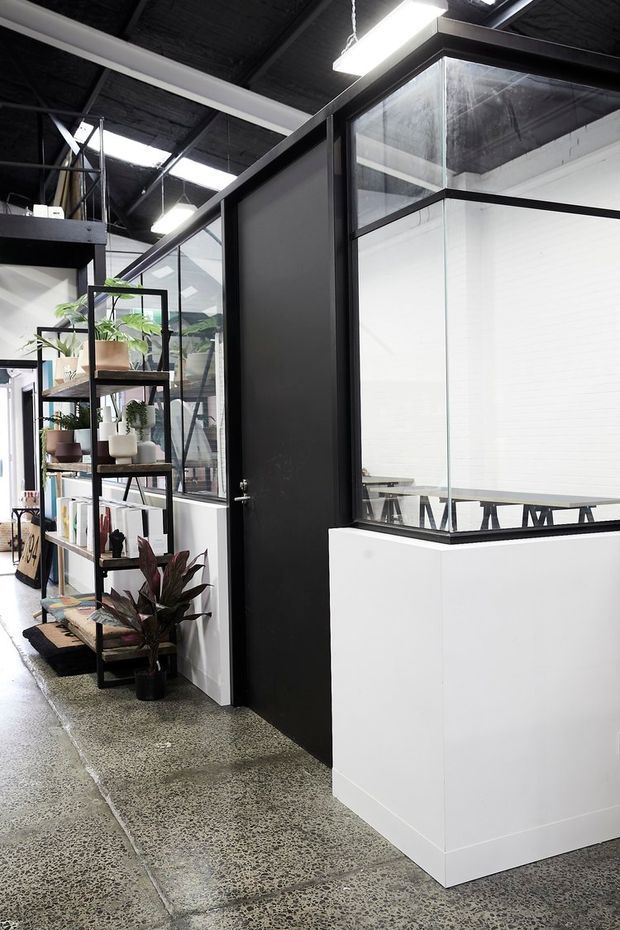 An industrial-style commercial interior with black-framed glazed office partitions, a matte black door, exposed dark ceiling structure, linear fluorescent lighting, and polished concrete flooring. A freestanding black metal shelving unit displays potted plants and small objects beside the circulation area. White partition walls and a meeting table are visible through the glazing.